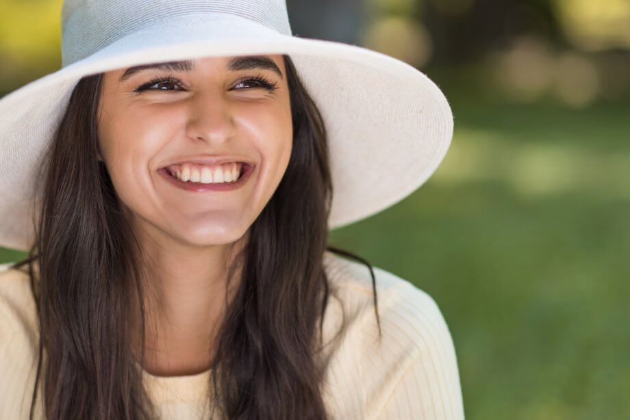 Close up photograph of a woman with long brown hair, smiling while wearing a white hat. Trees can be seen in the background.