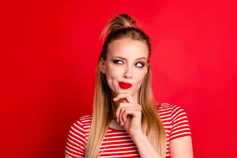 Photograph of a woman with red lipstick thinking about something. She has long hair, standing in front of a red background.